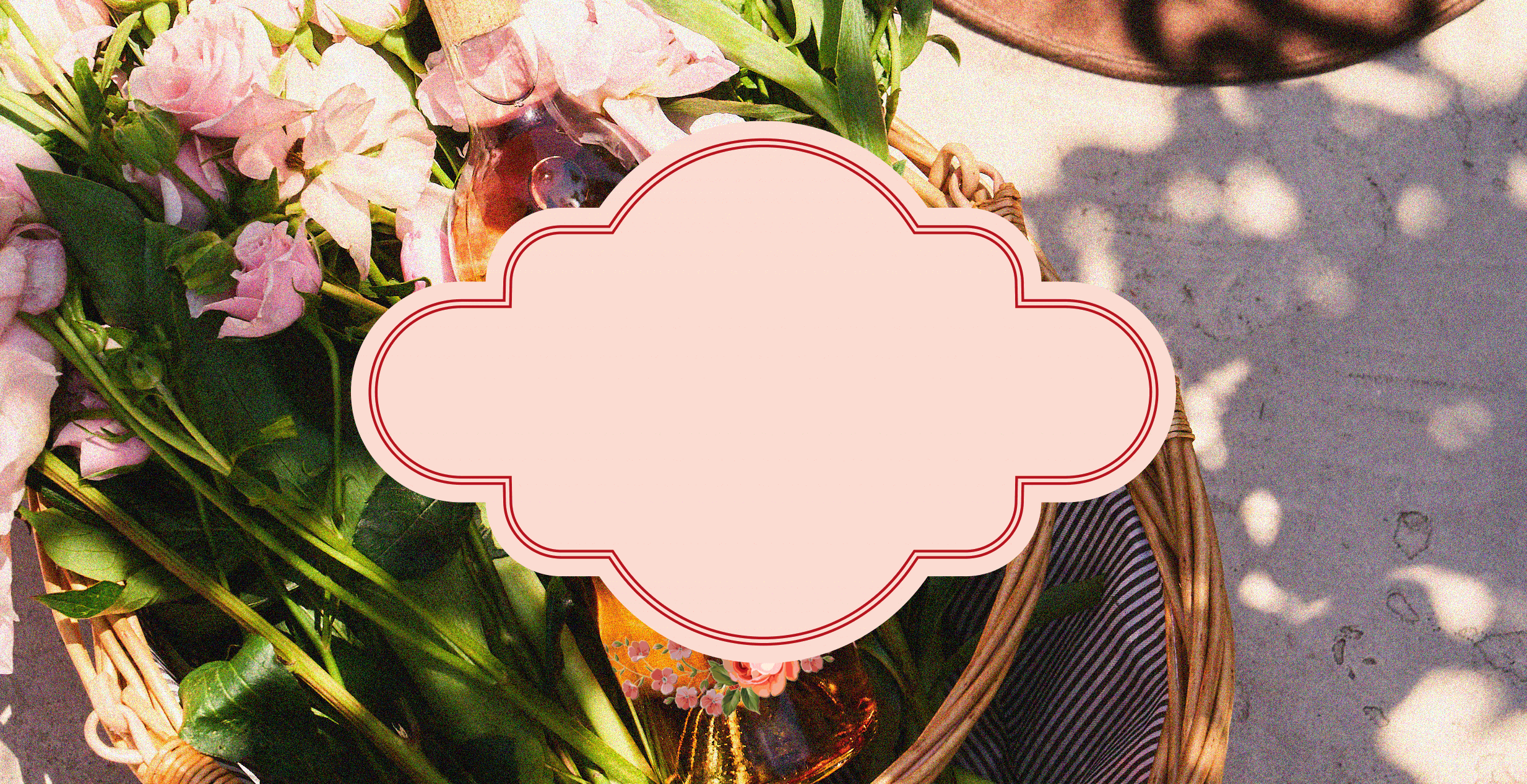 A wicker basket filled with pink flowers, greenery, and a bottle of rosé sits on the ground under dappled sunlight. At the center, a decorative pink label frame with a red outline is placed, leaving space for custom text.