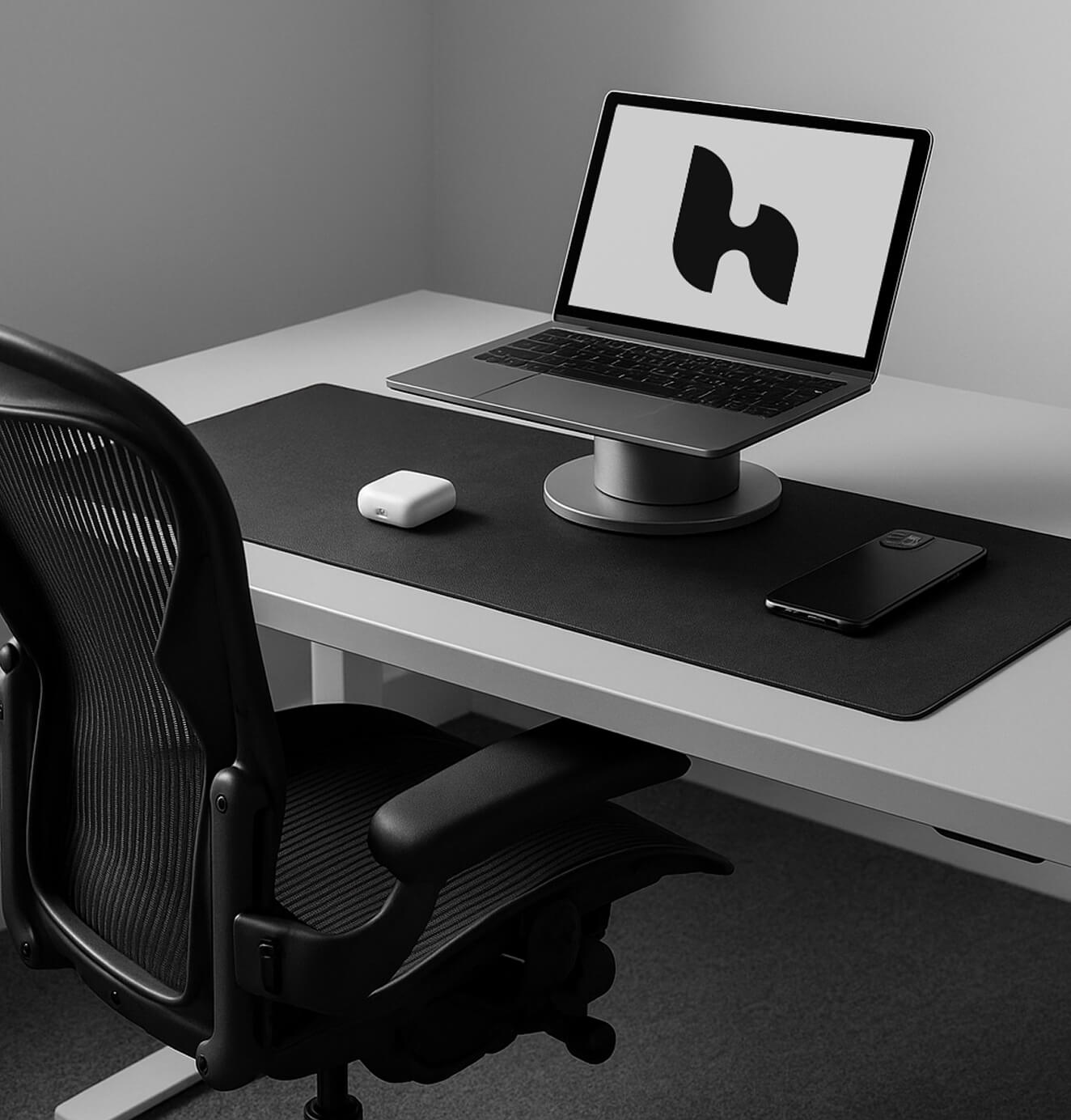 Minimalist black-and-white office setup featuring a laptop on a stand displaying a modern abstract logo, with a black desk mat, wireless earbuds, and smartphone neatly arranged.