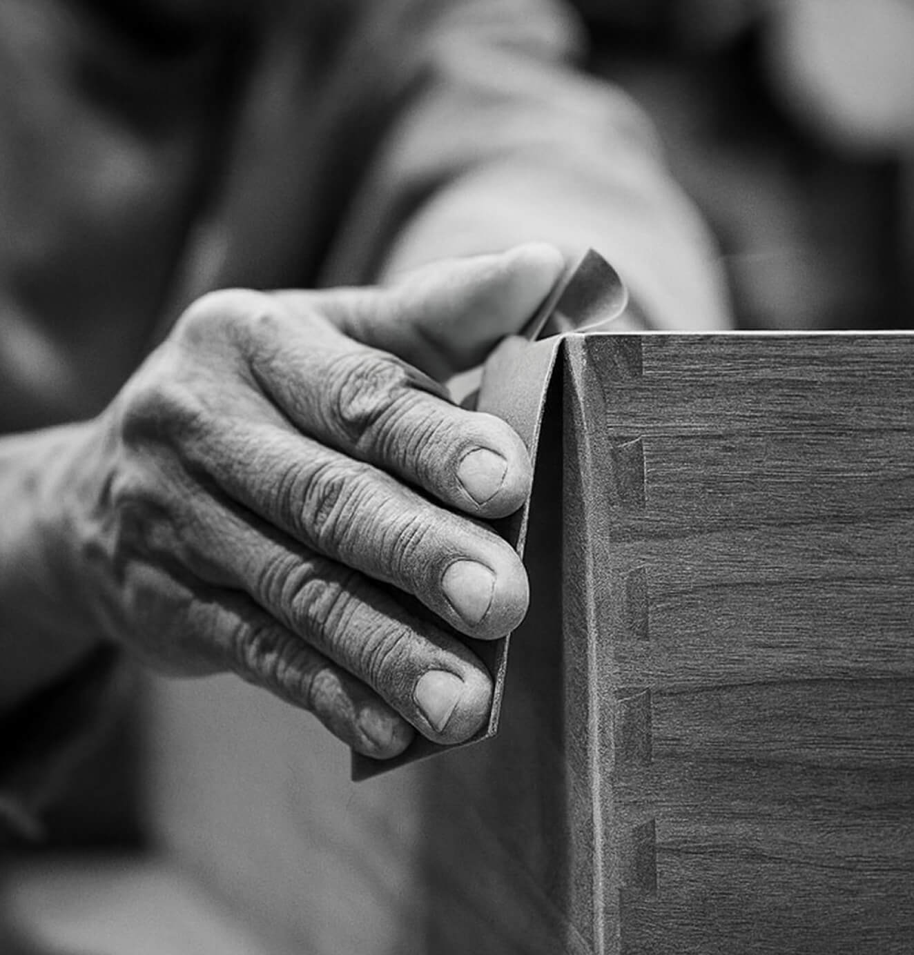 Close-up black-and-white photo of skilled hands sanding the corner of a wooden piece, highlighting craftsmanship and attention to detail.
