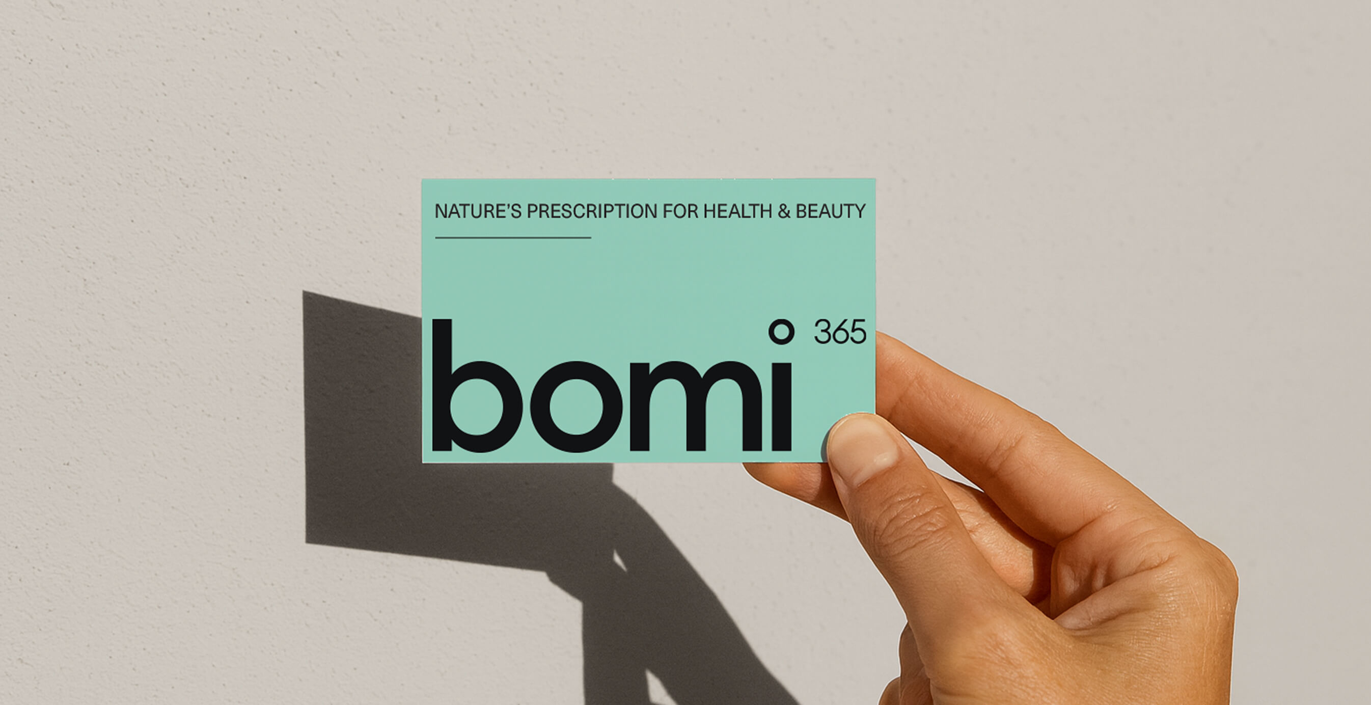 A hand holds a teal Bomi 365 card that reads “Nature’s Prescription for Health & Beauty” against a neutral wall.