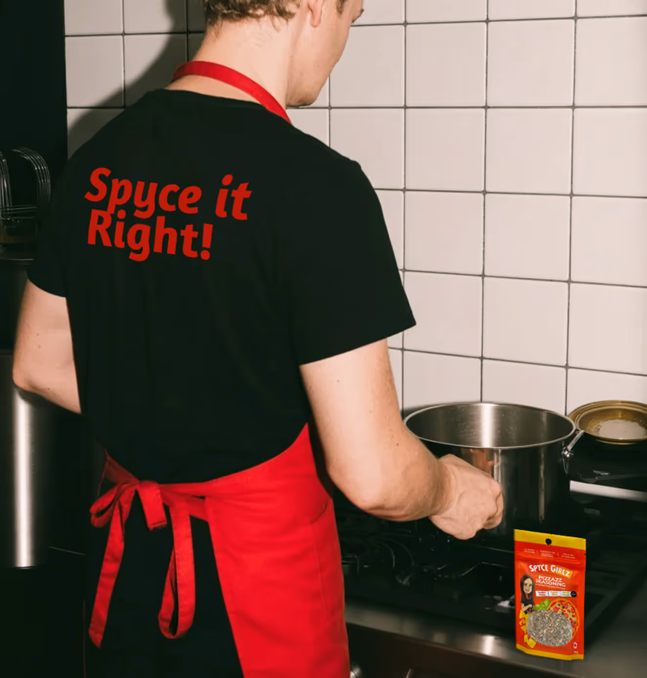 Man cooking in a kitchen, wearing a black t-shirt with red text “Spyce it Right!” and a red apron, with Spyce Girlz spice blend packaging placed in the scene.