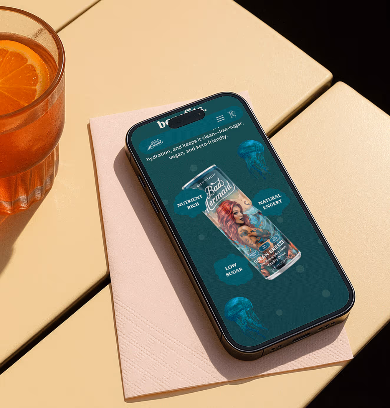 Animated GIF showing a smartphone on a café table next to a glass of orange drink. The phone screen displays a Bad Mermaid Ocean Breeze can with highlighted benefits such as nutrient-rich, low sugar, vegan, and natural energy.