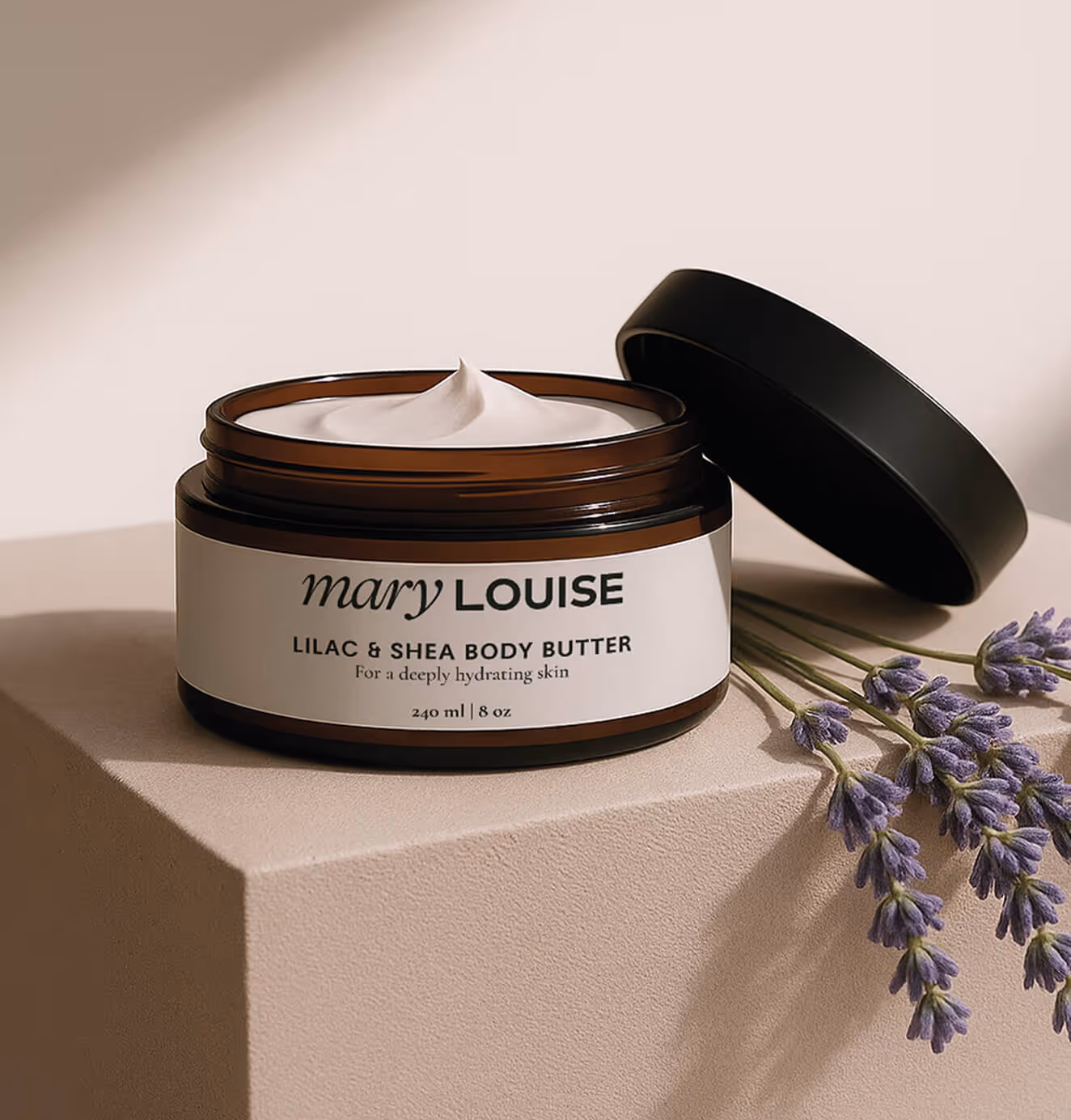 A jar of Mary Louise Lilac & Shea Body Butter with the lid open, showing creamy texture, placed on a beige surface beside sprigs of lavender.