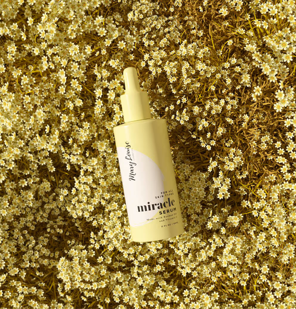 A Mary Louise Miracle Serum bottle with a dropper cap, lying on a bed of small yellow flowers.