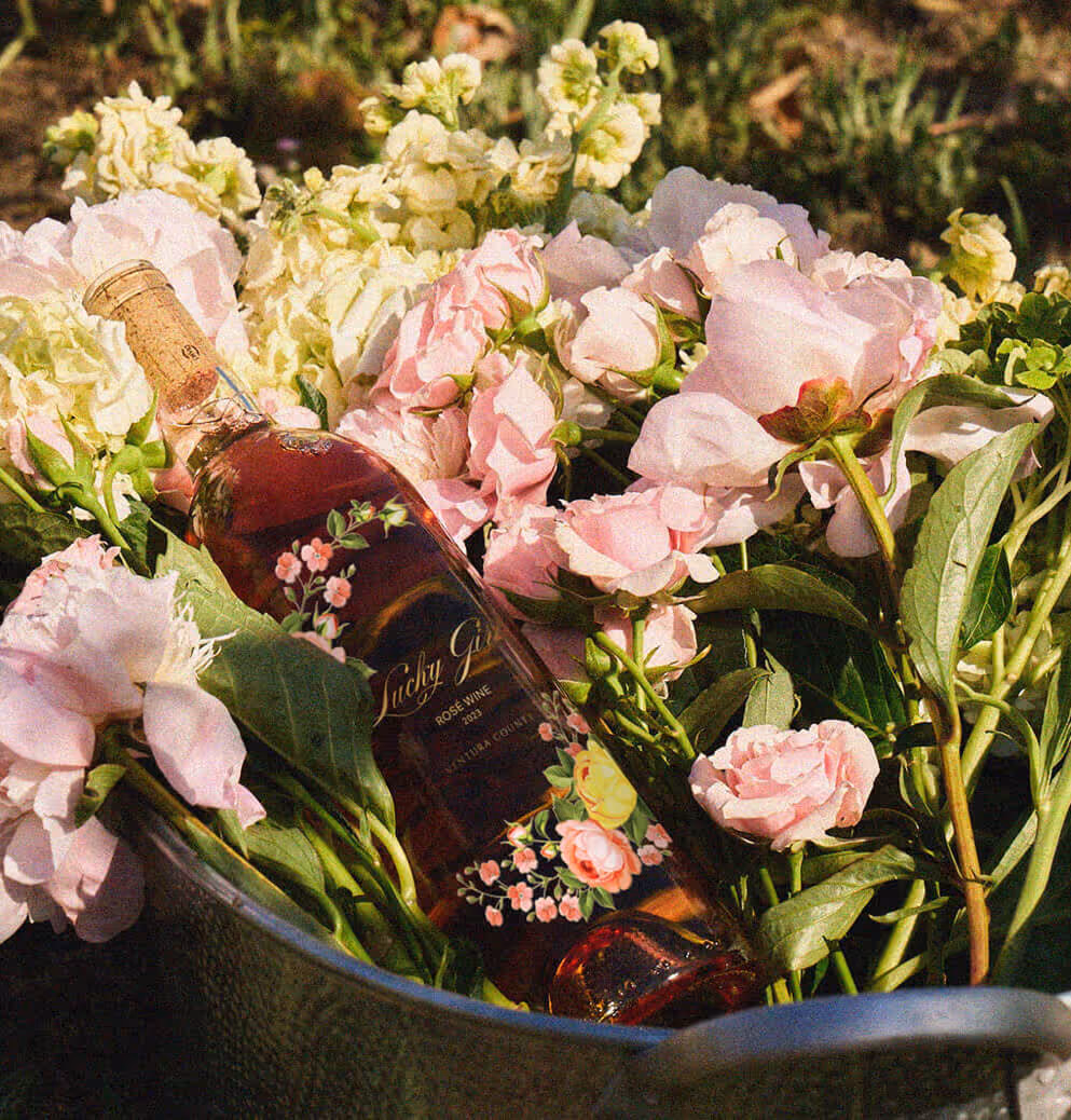A bottle of Lucky Girl rosé wine nestled among pink and white flowers in a rustic outdoor setting.