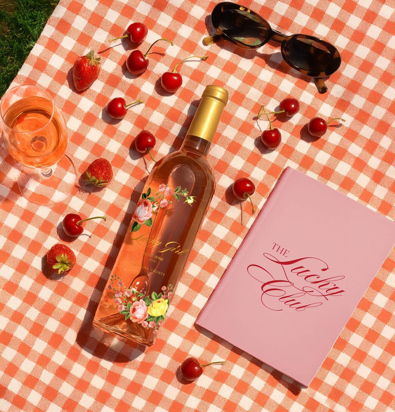 Lucky Girl rosé wine on a red-and-white checkered picnic blanket with cherries, strawberries, sunglasses, and a pink notebook titled The Lucky Club.