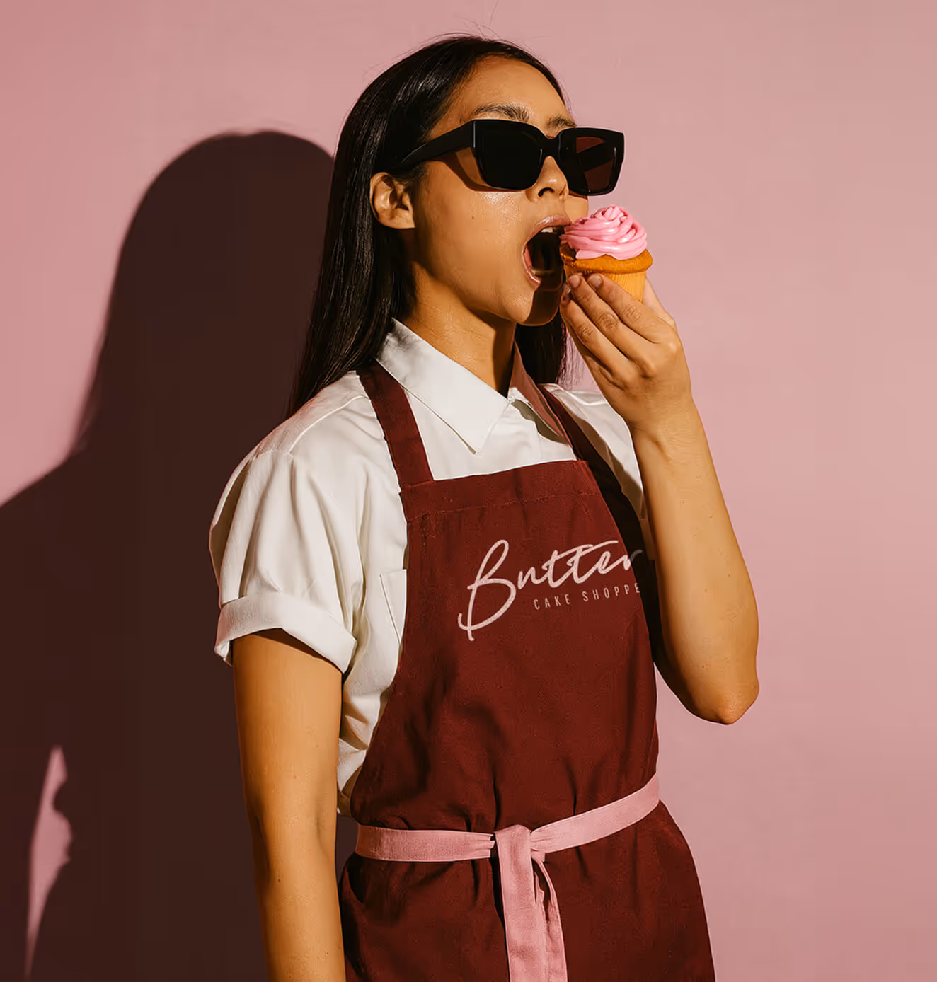 Woman wearing sunglasses and a burgundy apron with "Butter Cake Shoppe" logo takes a bite of a pink frosted cupcake against a pink background.