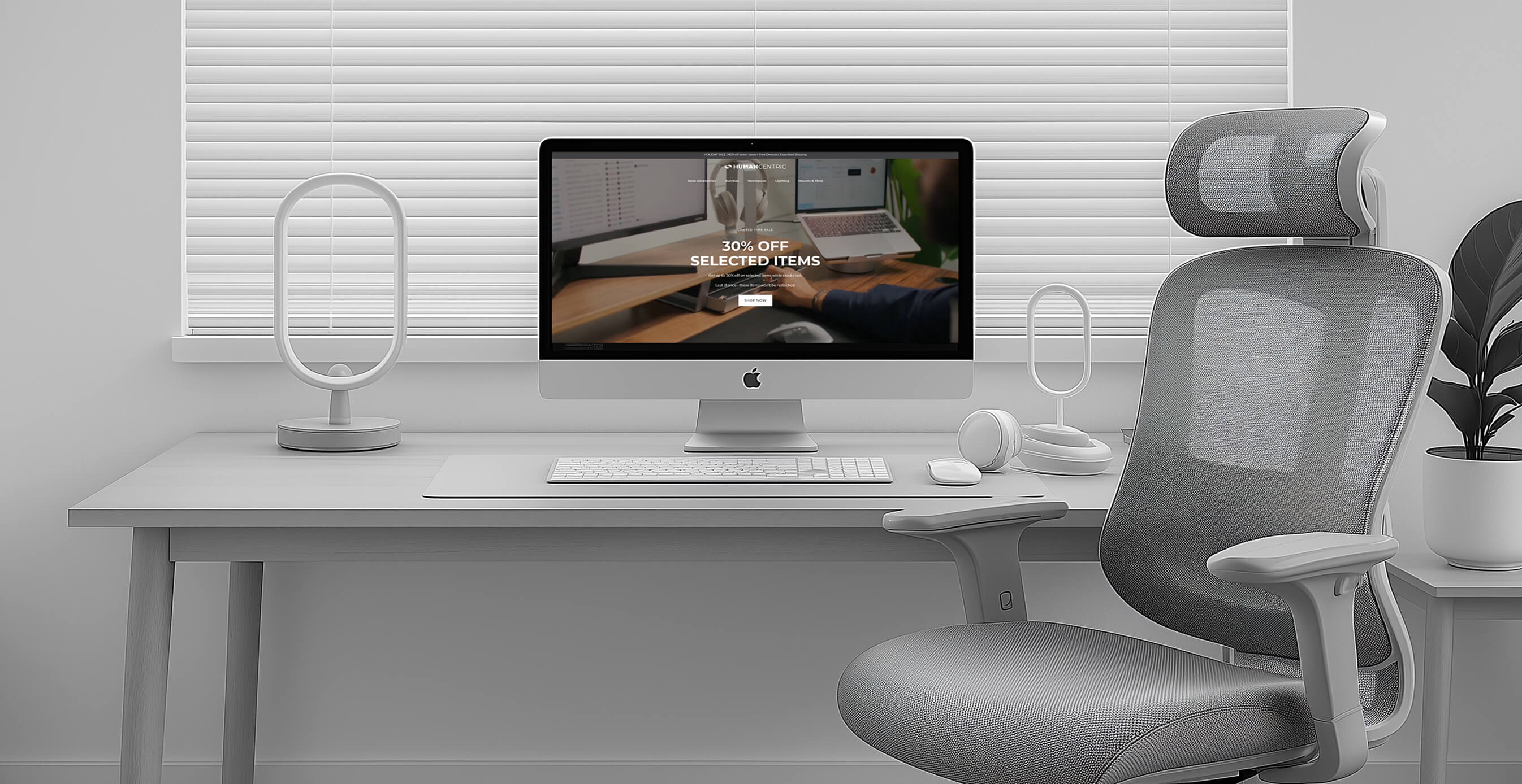 A minimalist home office setup with a sleek iMac on a white desk, modern accessories, and an ergonomic gray office chair in front of closed blinds.