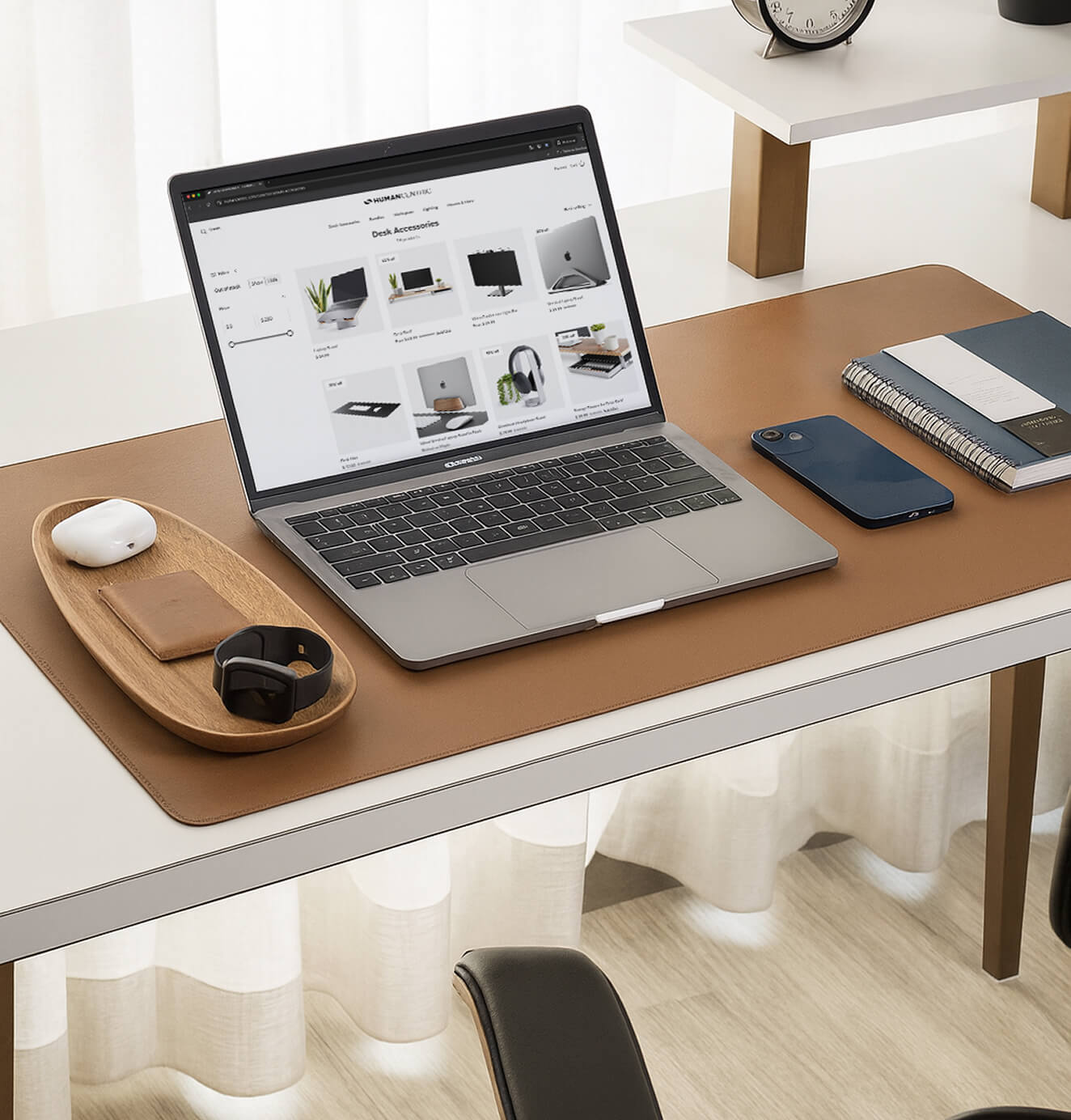 A close-up of a tidy desk with a laptop displaying an online store, surrounded by a phone, notebook, AirPods, smartwatch, and wooden tray on a leather desk mat.