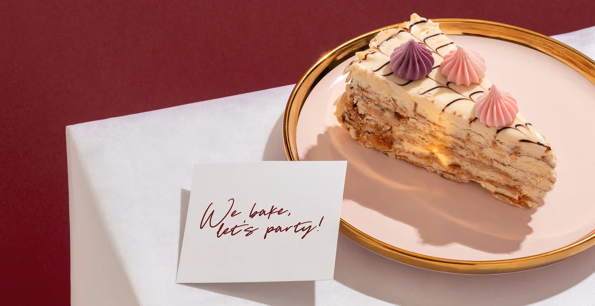 Close-up of a slice of layered cake with cream filling and pink and purple frosting dollops on a pink plate beside a card reading “We bake, let’s party!”