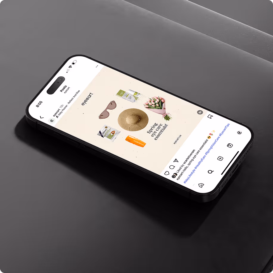 A black smartphone resting on a dark, textured surface displays an Instagram post by @eyebuydirect. The post features a flat lay image with summer-themed items including sunglasses, a straw sunhat, a bouquet of flowers, two boxes of eyeglasses, and a bottl