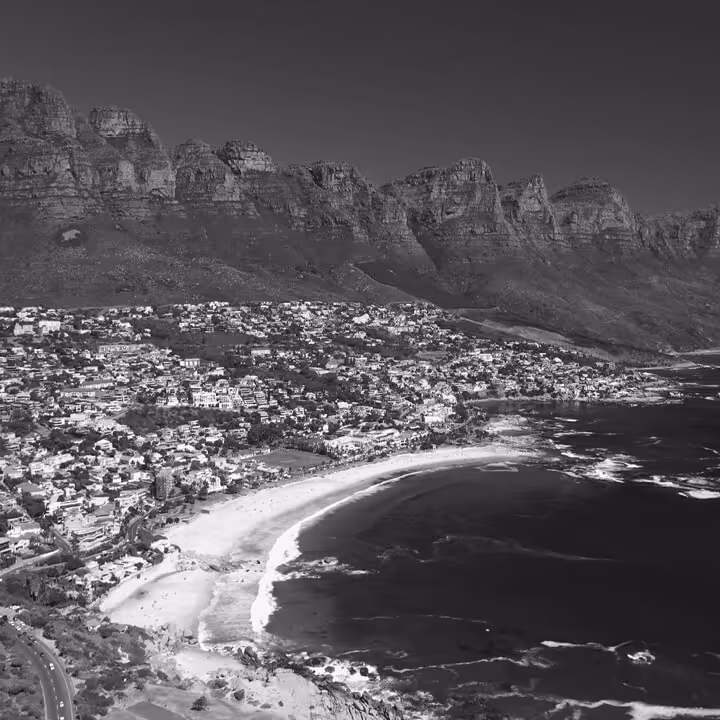 Black-and-white aerial view of Cape Town’s coastline with sandy beaches, residential areas, and the rugged peaks of the Twelve Apostles mountain range.