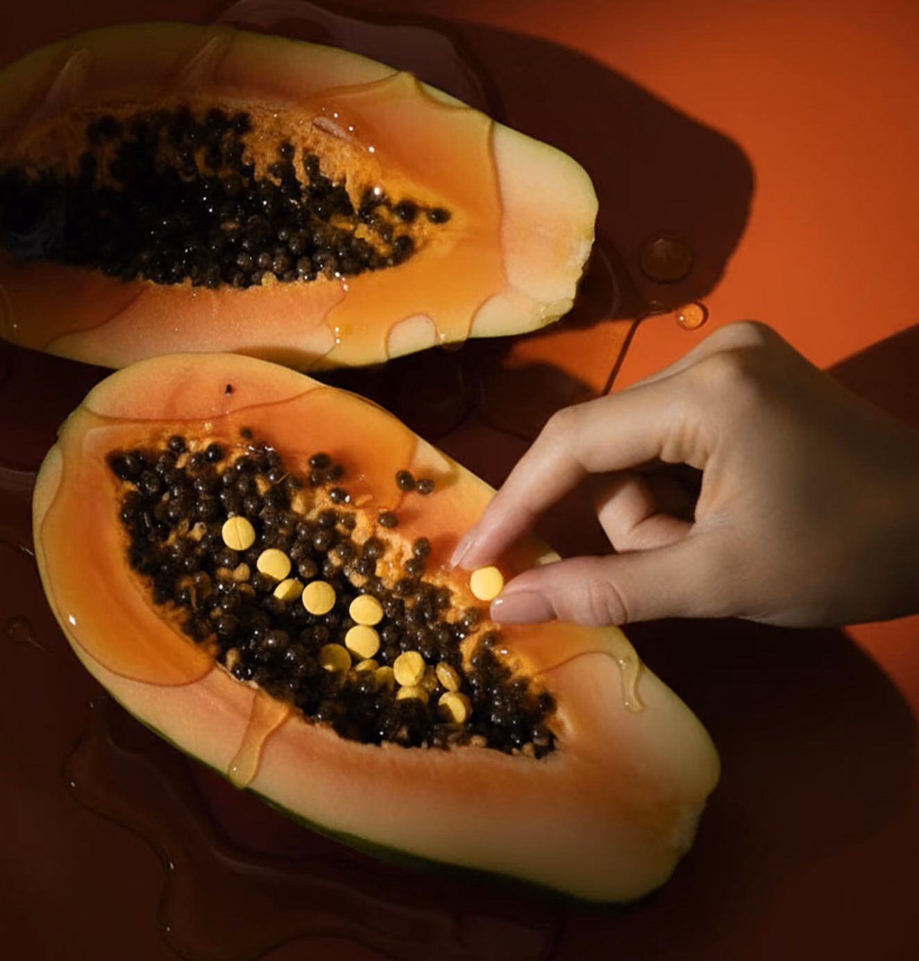 Close-up of sliced papaya drizzled with liquid, filled with black seeds and yellow supplement tablets, as a hand places one tablet into the fruit.