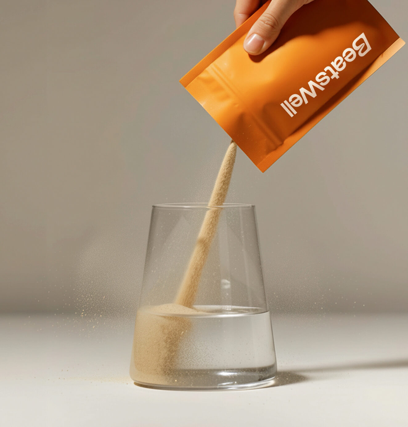 Hand pouring beige powdered supplement from an orange BeatsWell sachet into a clear glass of water, with fine particles visible as they dissolve.