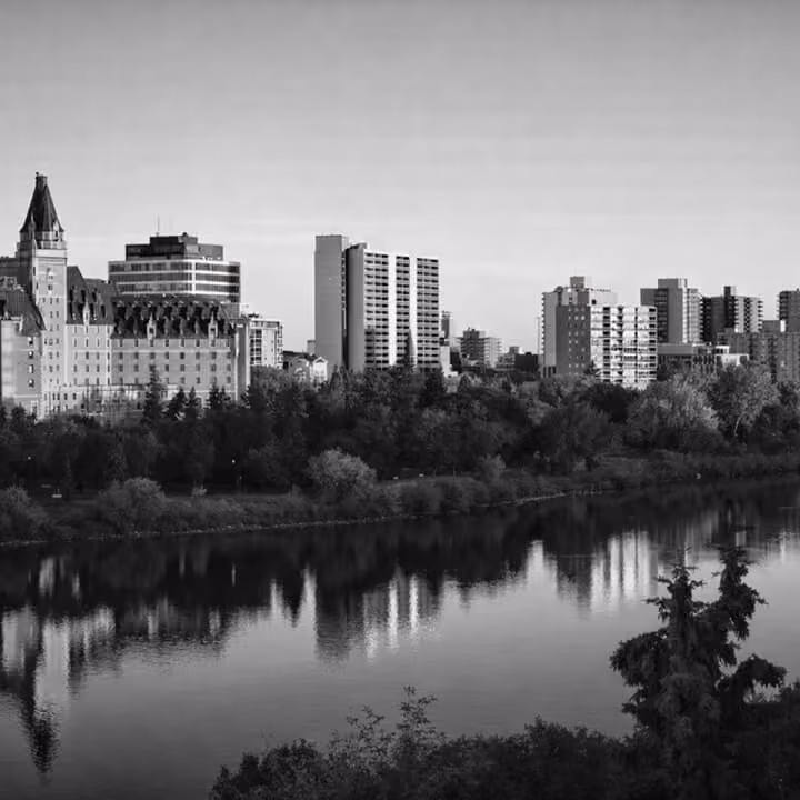 Black and white view of Saskatoon skyline along the South Saskatchewan River, featuring historic architecture and waterfront reflections.