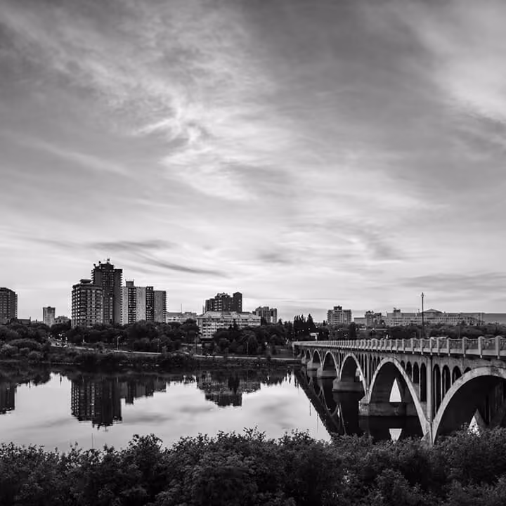 Black and white view of Saskatoon skyline and arched bridge over the South Saskatchewan River at sunset.