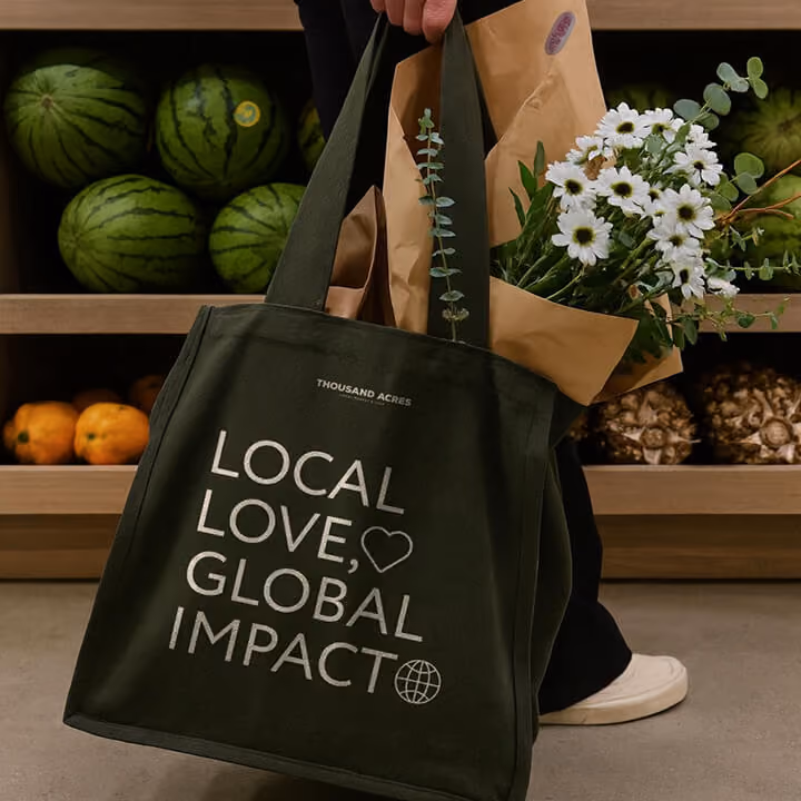 Branded reusable grocery tote reading “Local Love, Global Impact” filled with flowers and produce in a market setting.