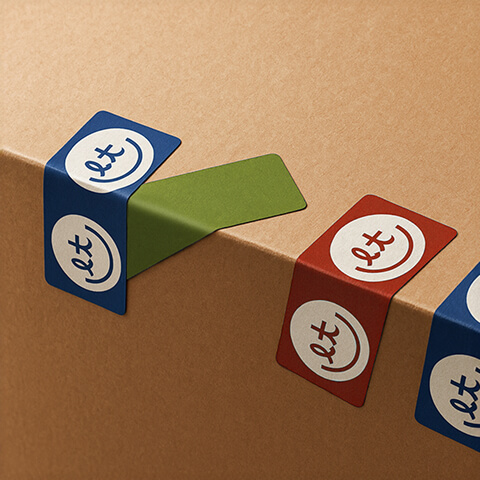 Close-up of branded adhesive tabs in blue, green, and red wrapped over a cardboard box edge, featuring a minimalist circular logo.