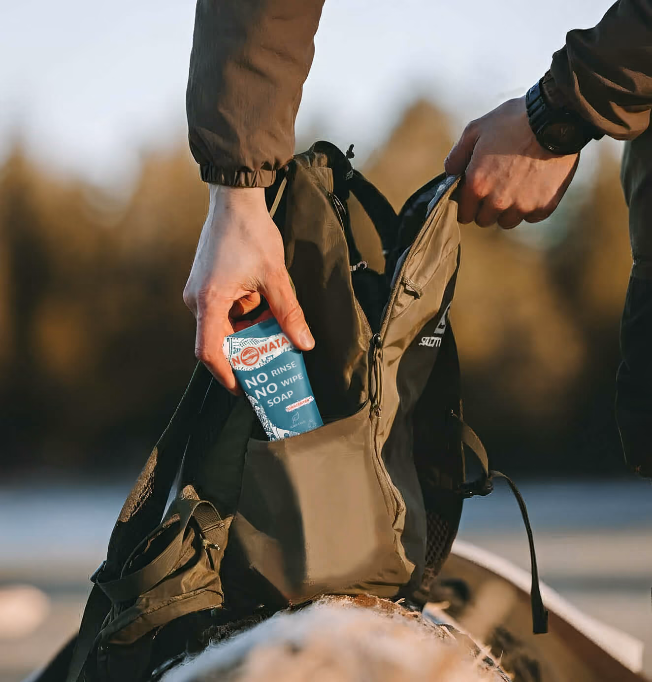 Person placing a tube of NOWATA no-rinse soap into a backpack outdoors near water at sunset.