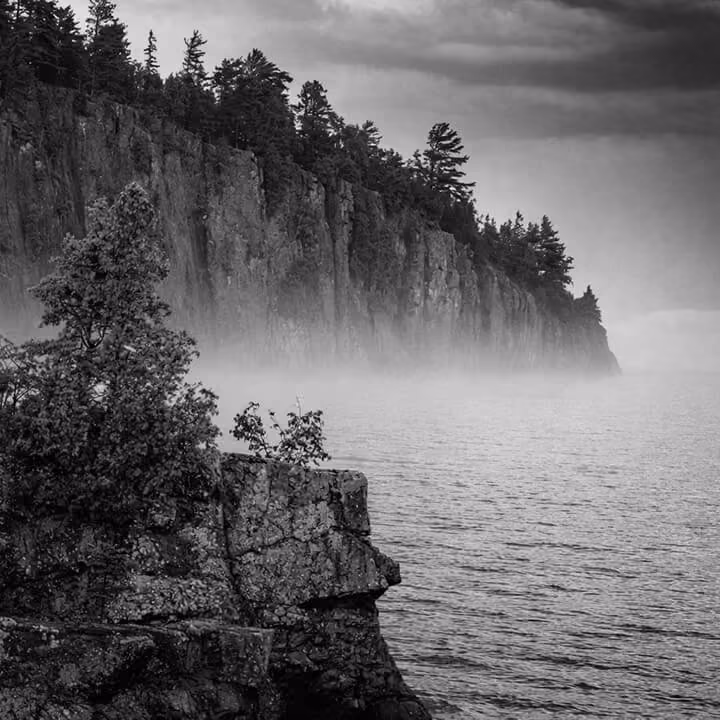 Black and white landscape of rugged cliffs along Lake Superior in Minnesota with pine trees and mist rising over the water.