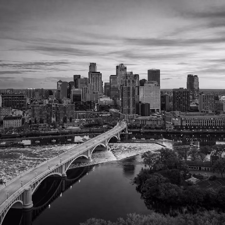 Black and white aerial view of the Minneapolis skyline with a large bridge crossing the Mississippi River.