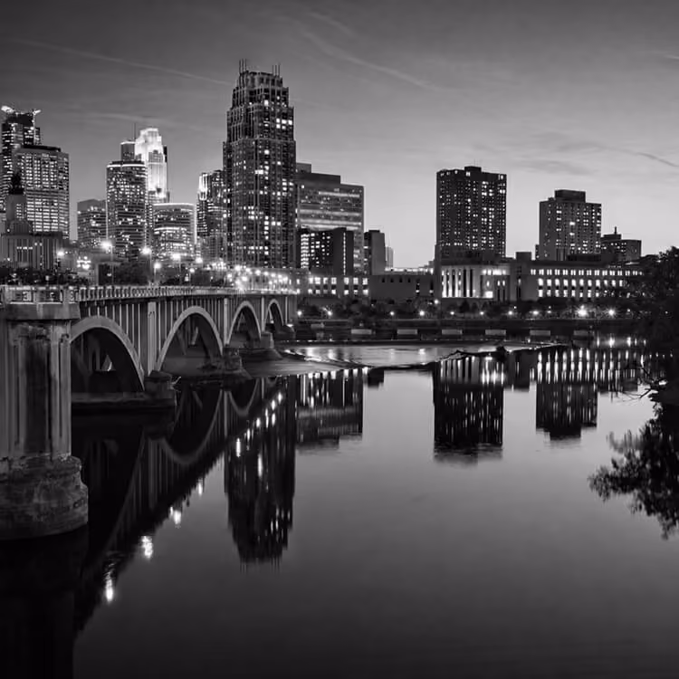 Black and white view of the Minneapolis skyline at night with an illuminated bridge reflecting on the Mississippi River.