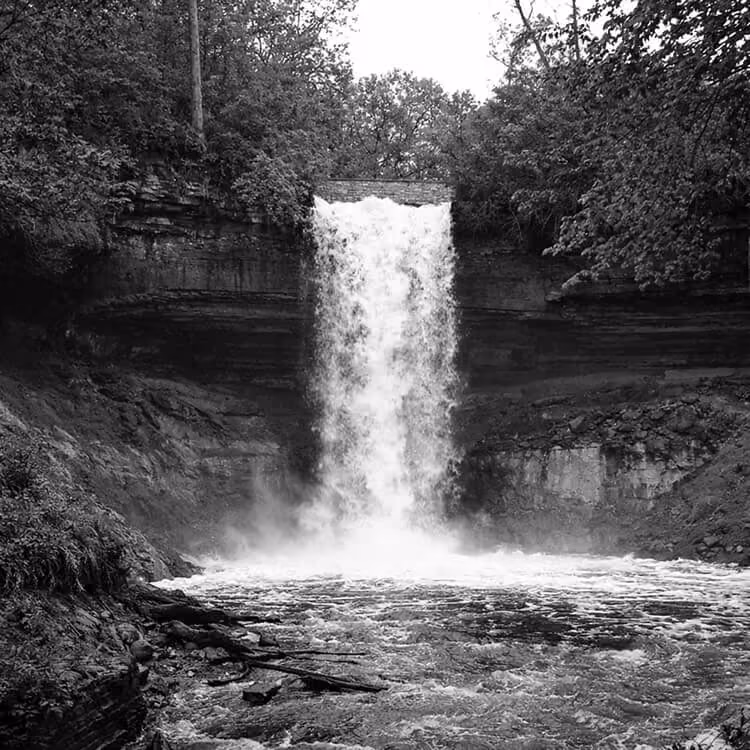 Black and white photograph of Minnehaha Falls cascading over rocky cliffs surrounded by trees in Minneapolis.
