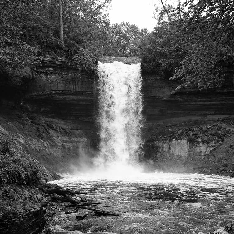 Black and white photograph of Minnehaha Falls cascading over rocky cliffs surrounded by trees in Minneapolis.