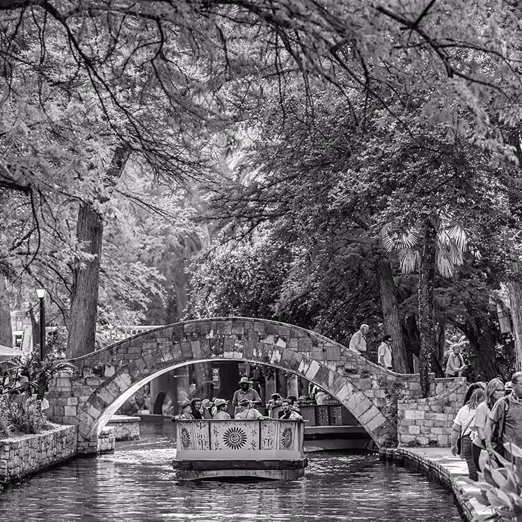 Black and white image of a tour boat passing under a stone bridge along the San Antonio River Walk surrounded by trees and pedestrians.