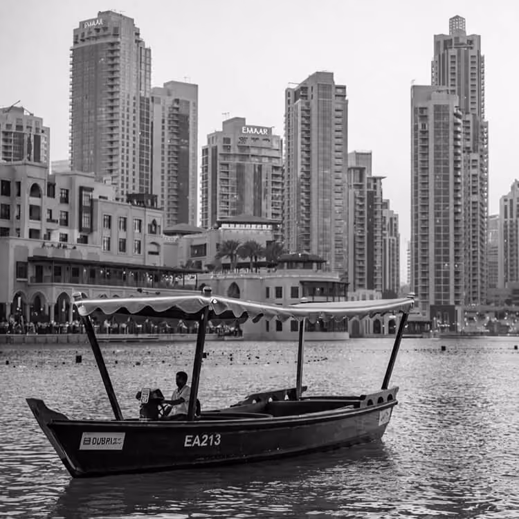 Black and white image of a traditional boat on the water with Dubai Marina skyscrapers in the background.