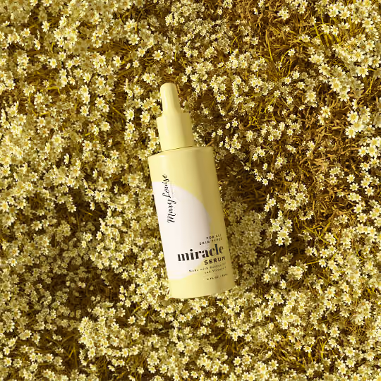 Yellow Miracle Face Serum bottle placed on a bed of small chamomile flowers in a bright botanical skincare scene.