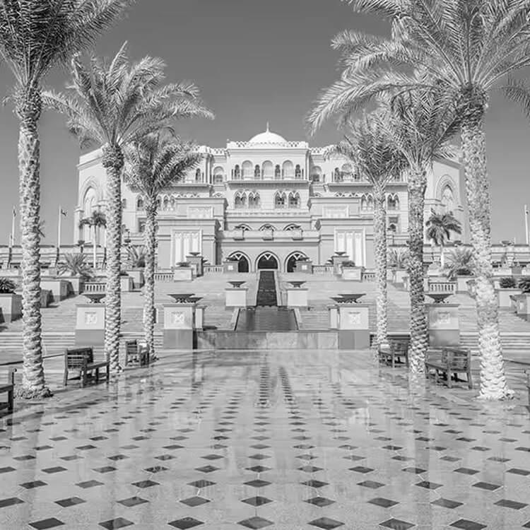 Black and white front view of Emirates Palace hotel framed by palm trees and a patterned courtyard in Abu Dhabi.