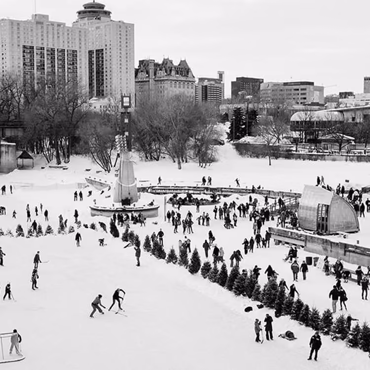 Black and white image of people ice skating at The Forks in Winnipeg during winter with city buildings in the background.