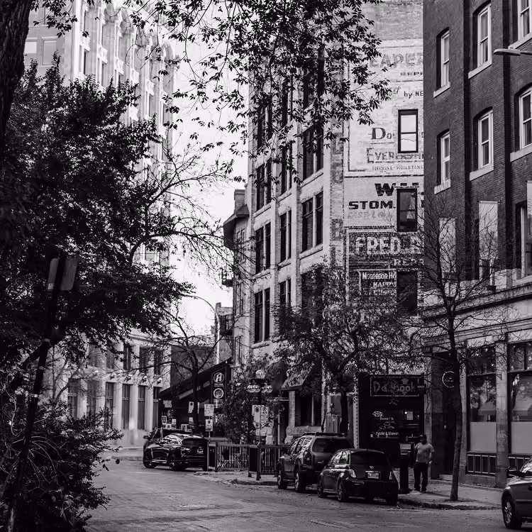 Black and white view of a historic street in Winnipeg’s Exchange District with brick buildings, parked cars, and trees.