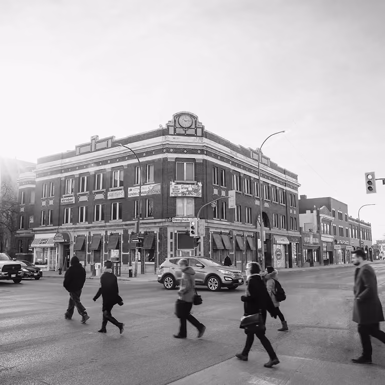 Black and white photo of pedestrians crossing a downtown Winnipeg street near a historic corner building.