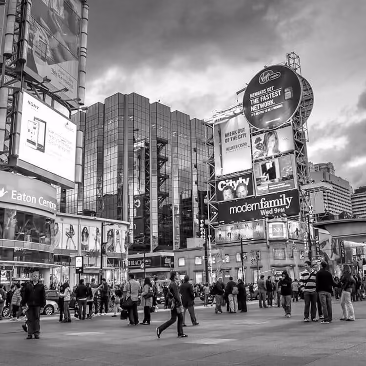 Black and white image of people crossing the street at Yonge-Dundas Square surrounded by digital billboards in downtown Toronto.