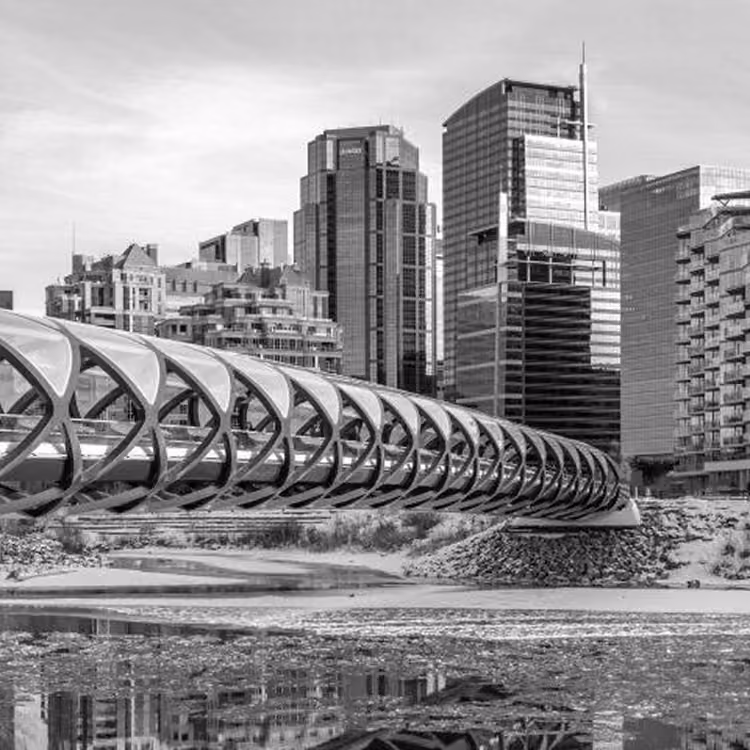 Black and white image of the Peace Bridge spanning the Bow River with Calgary’s downtown skyline in the background.