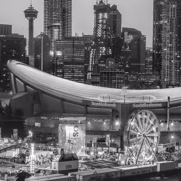 Black and white view of the Scotiabank Saddledome and Calgary Stampede grounds with a Ferris wheel and city skyline at night.