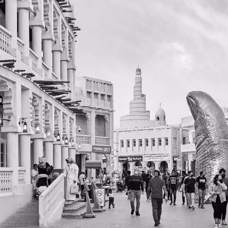 Black and white photo of people walking through Souq Waqif with traditional buildings and the Fanar Islamic Cultural Center in the background.