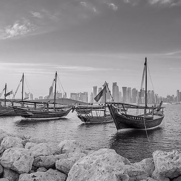 Black and white image of traditional wooden dhow boats along the Doha coastline with the city skyline in the background.