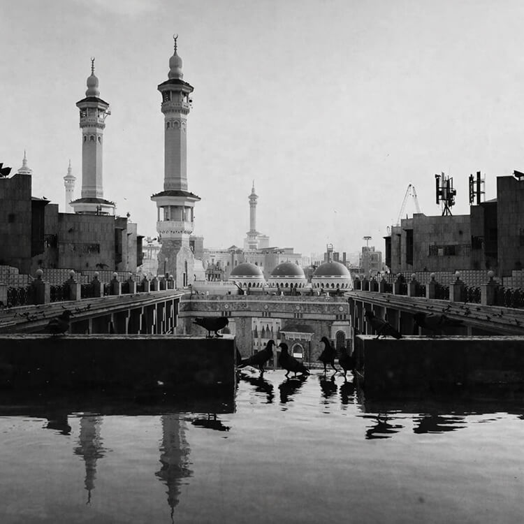 Black and white view of a mosque with tall minarets and reflective courtyard in a Saudi Arabian city.