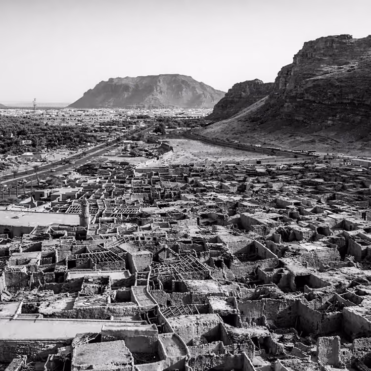 Black and white aerial view of a traditional village surrounded by mountains and desert terrain in Saudi Arabia.