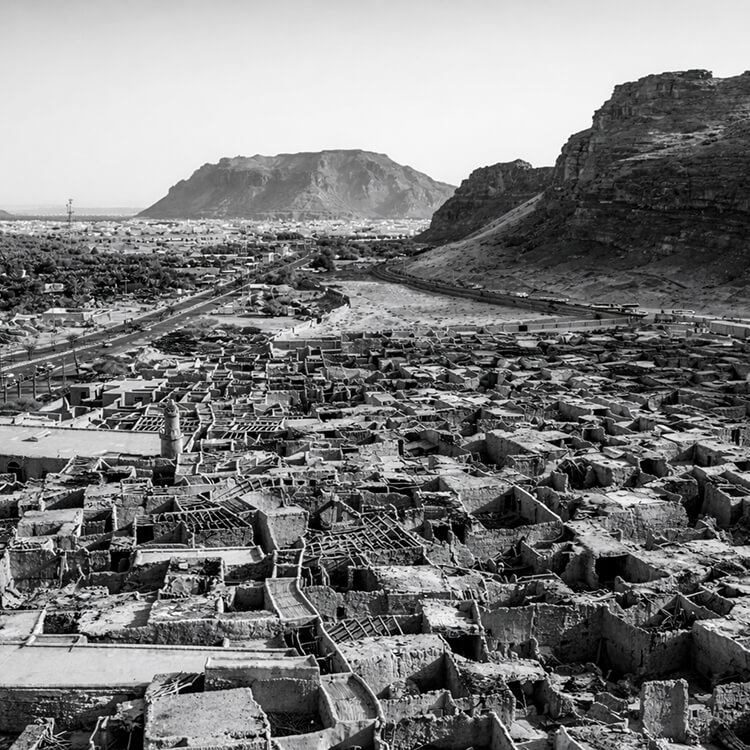 Black and white aerial view of a traditional village surrounded by mountains and desert terrain in Saudi Arabia.