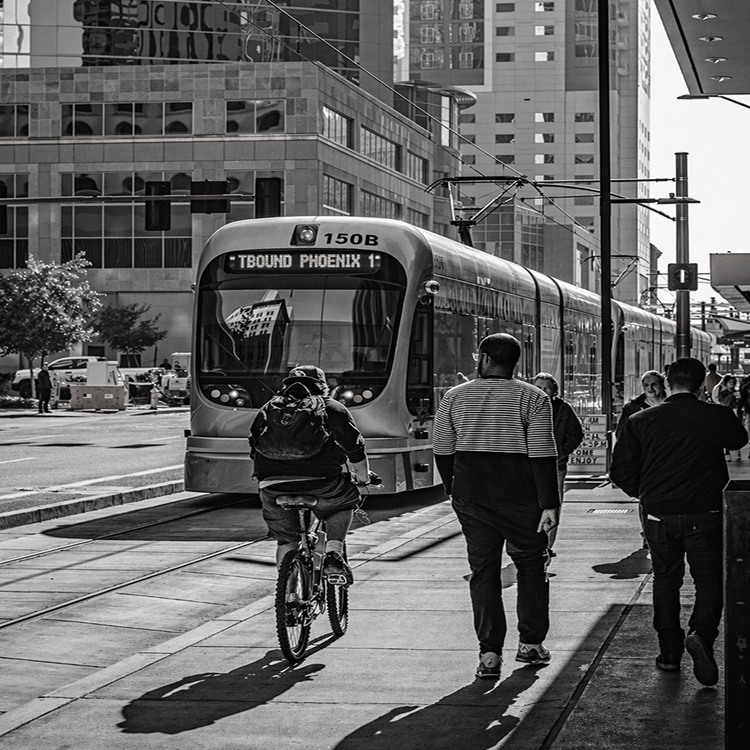 Black and white image of Phoenix light rail train with pedestrians and cyclist in an urban downtown setting.