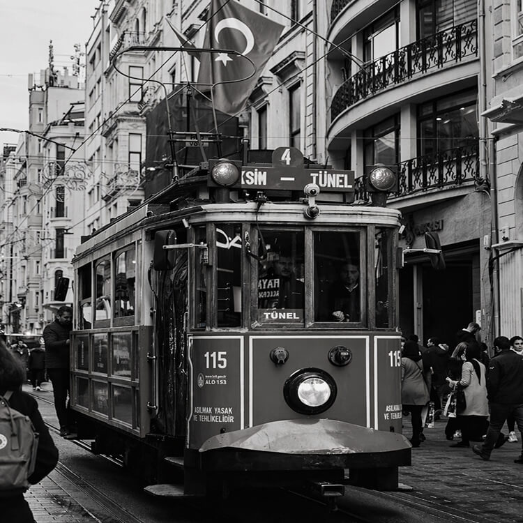 Black and white image of a vintage red tram on Istiklal Street with pedestrians and Turkish flags.