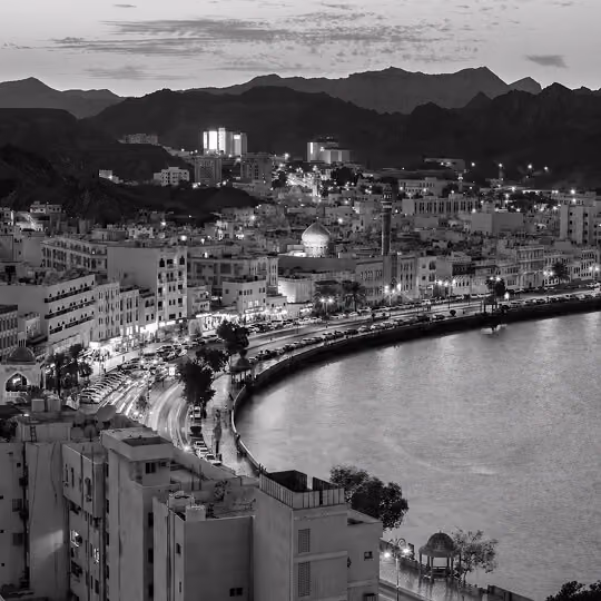 Aerial view of Muscat coastline with city lights, waterfront road, and surrounding mountains at dusk.