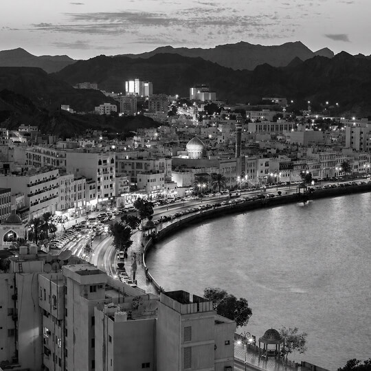 Aerial view of Muscat coastline with city lights, waterfront road, and surrounding mountains at dusk.