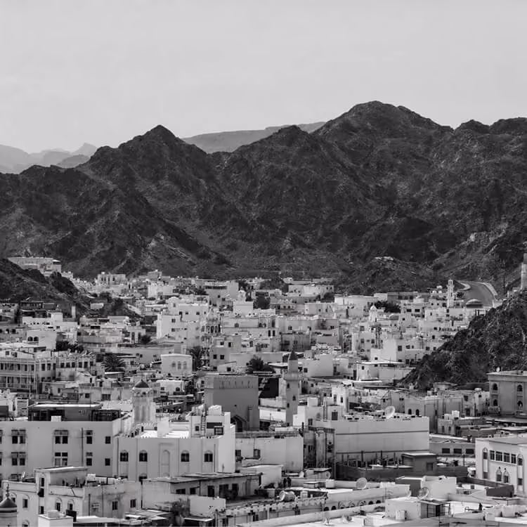 Dense Muscat cityscape with white buildings clustered beneath rugged mountain range in Oman.