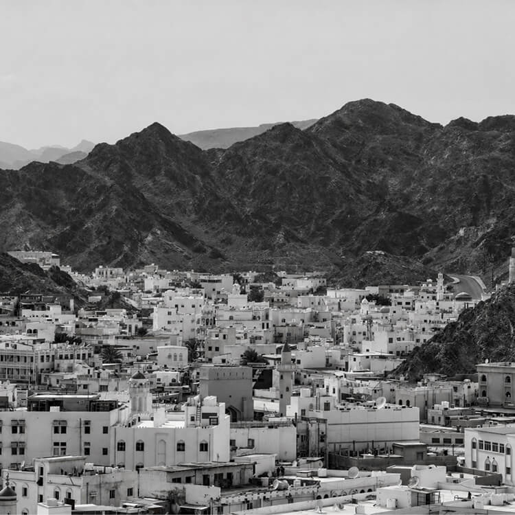 Dense Muscat cityscape with white buildings clustered beneath rugged mountain range in Oman.
