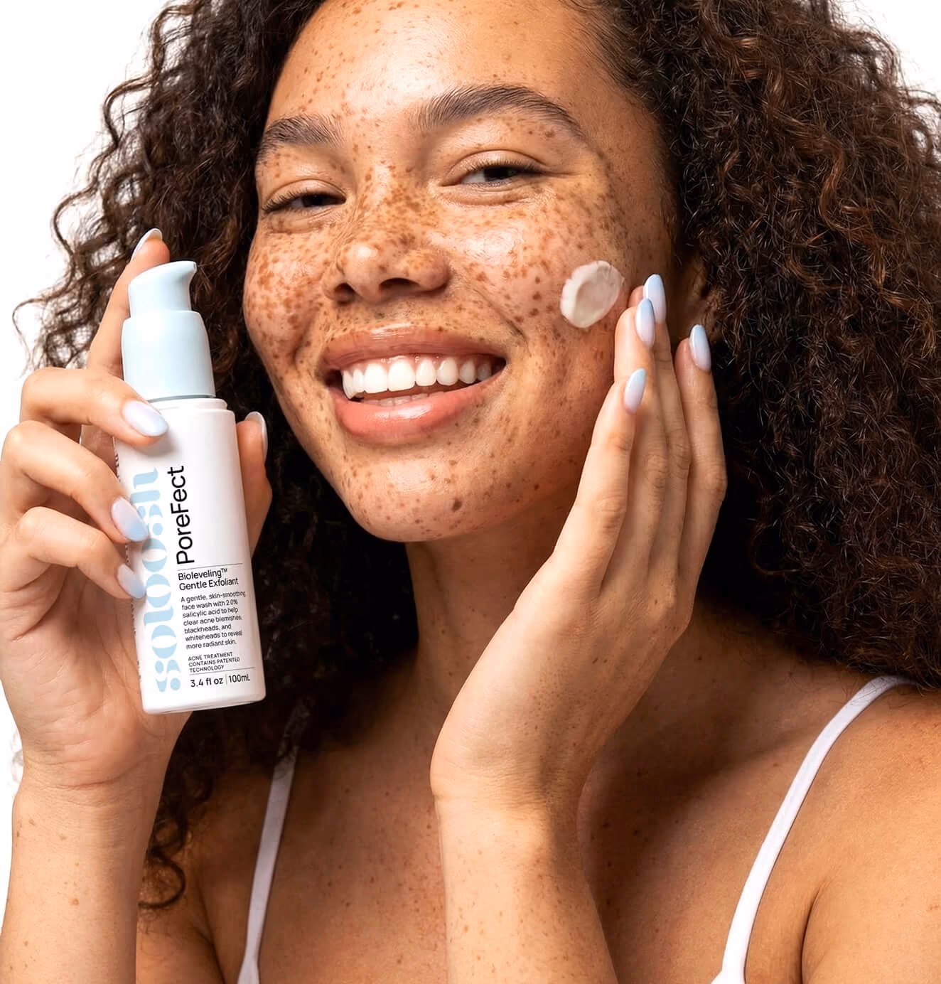 Smiling woman with freckles applying skincare cream while holding Soloosh Porefect exfoliant bottle.